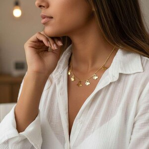 May include: A gold-colored necklace with butterfly pendants. The necklace is worn over a white button-down shirt. The image is a close-up of the necklace and the person wearing it. The background is blurred, with a warm, neutral color palette.