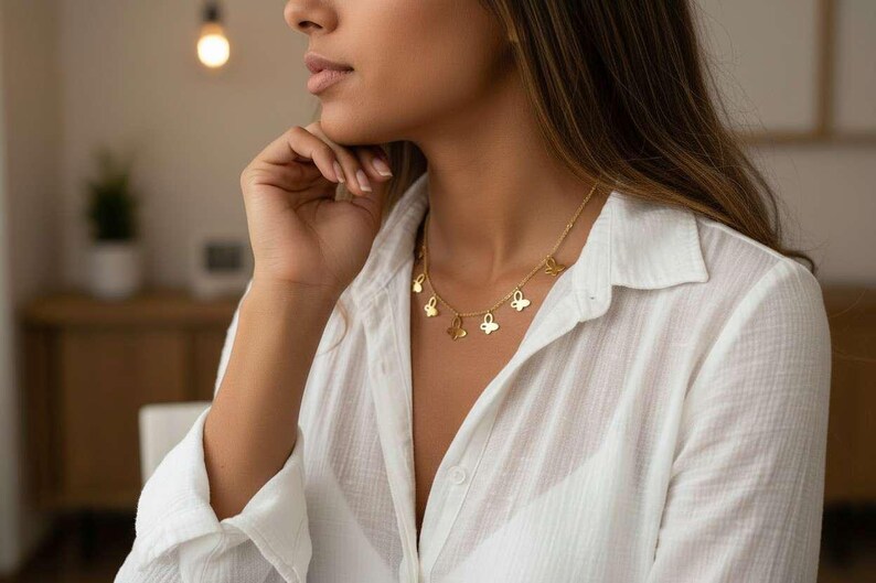 May include: A gold-colored necklace with butterfly pendants. The necklace is worn over a white button-down shirt. The image is a close-up of the necklace and the person wearing it. The background is blurred, with a warm, neutral color palette.
