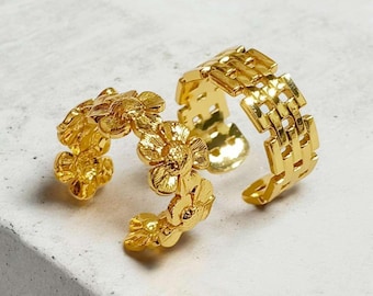 May include: Two gold-colored rings. One ring features a floral design, with multiple small flowers. The other ring has a woven, rectangular pattern. Both rings are open-ended, allowing for adjustable sizing.