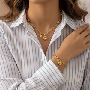 Puede incluir: Un conjunto de collar y pulsera dorados. El collar y la pulsera presentan un diseño de eslabones de cadena con un dije en forma de corazón y un pequeño dije redondo. La modelo lleva una camisa abotonada a rayas blancas y azules.