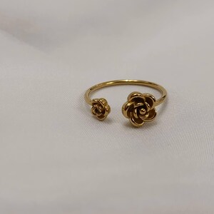 May include: A gold-colored ring featuring two open rose flower designs. The ring has a delicate band and a floral motif, with one rose on each end of the open design. The ring is set against a white background.