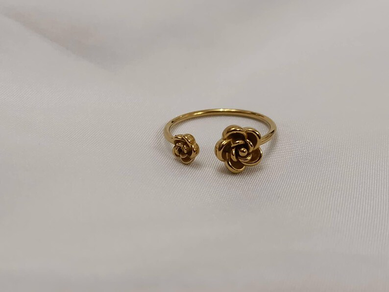 May include: A gold-colored ring featuring two open rose flower designs. The ring has a delicate band and a floral motif, with one rose on each end of the open design. The ring is set against a white background.