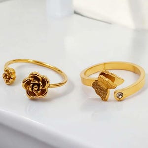 May include: Two gold-colored rings. One ring features a rose design, with a small rose on one end and a larger rose on the other. The second ring has a butterfly design with a small clear gem. Both rings are open-ended.