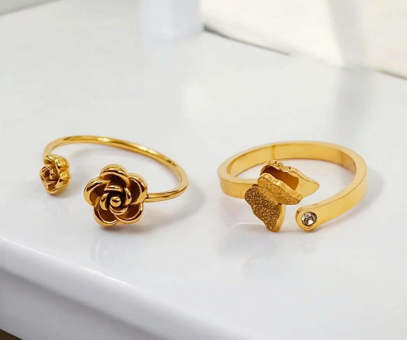 May include: Two gold-colored rings. One ring features a rose design, with a small rose on one end and a larger rose on the other. The second ring has a butterfly design with a small clear gem. Both rings are open-ended.