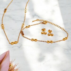 Puede incluir: Un conjunto de joyas de color dorado con diseños de símbolos de infinito. El conjunto incluye un collar, una pulsera y un par de pendientes. Las joyas se exhiben sobre una superficie de mármol blanco.