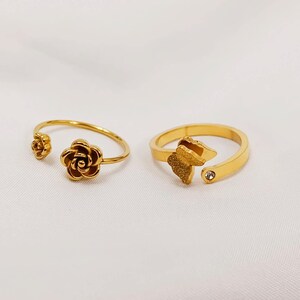 May include: Two gold-colored rings. One ring features a rose design, while the other has a butterfly design with a small, clear gemstone. Both rings have an open design, allowing for size adjustment. The rings are displayed on a white surface.