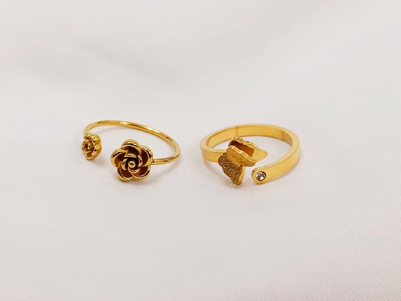 May include: Two gold-colored rings. One ring features a rose design, while the other has a butterfly design with a small, clear gemstone. Both rings have an open design, allowing for size adjustment. The rings are displayed on a white surface.