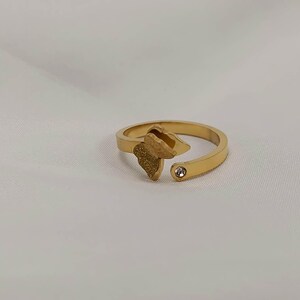 May include: A gold-colored ring with an open design. The ring features a textured leaf-shaped element and a small, clear gemstone. The ring is set against a white background.