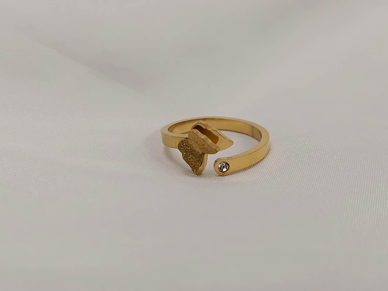 May include: A gold-colored ring with an open design. The ring features a textured leaf-shaped element and a small, clear gemstone. The ring is set against a white background.