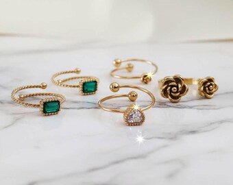 May include: A collection of five gold-toned rings featuring various designs, including two rings with rectangular green stones, one ring with a heart-shaped clear stone, one ring with two small spheres, and one ring with two sculpted rose-shaped accents.