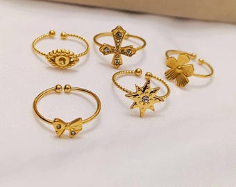 May include: Five gold-colored adjustable rings with various designs. The rings feature a bow, a flower, a cross, a starburst, and a circular design. Each ring has a delicate, thin band and is set against a white background.