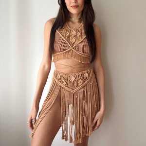 May include: Tan macrame halter top and skirt set. The top features a geometric design with fringe, and the skirt has a high slit and dangling fringe. The set is made of woven rope.