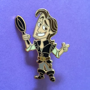May include: Gold-colored enamel pin featuring a cartoon character holding a frying pan. The character has brown hair, a black vest, and a light green shirt. The character is smiling and pointing with their right hand.