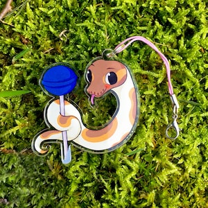 May include: A white and brown snake with a blue lollipop charm on a pink and white lanyard. The snake is curled up and has a cute cartoon face.