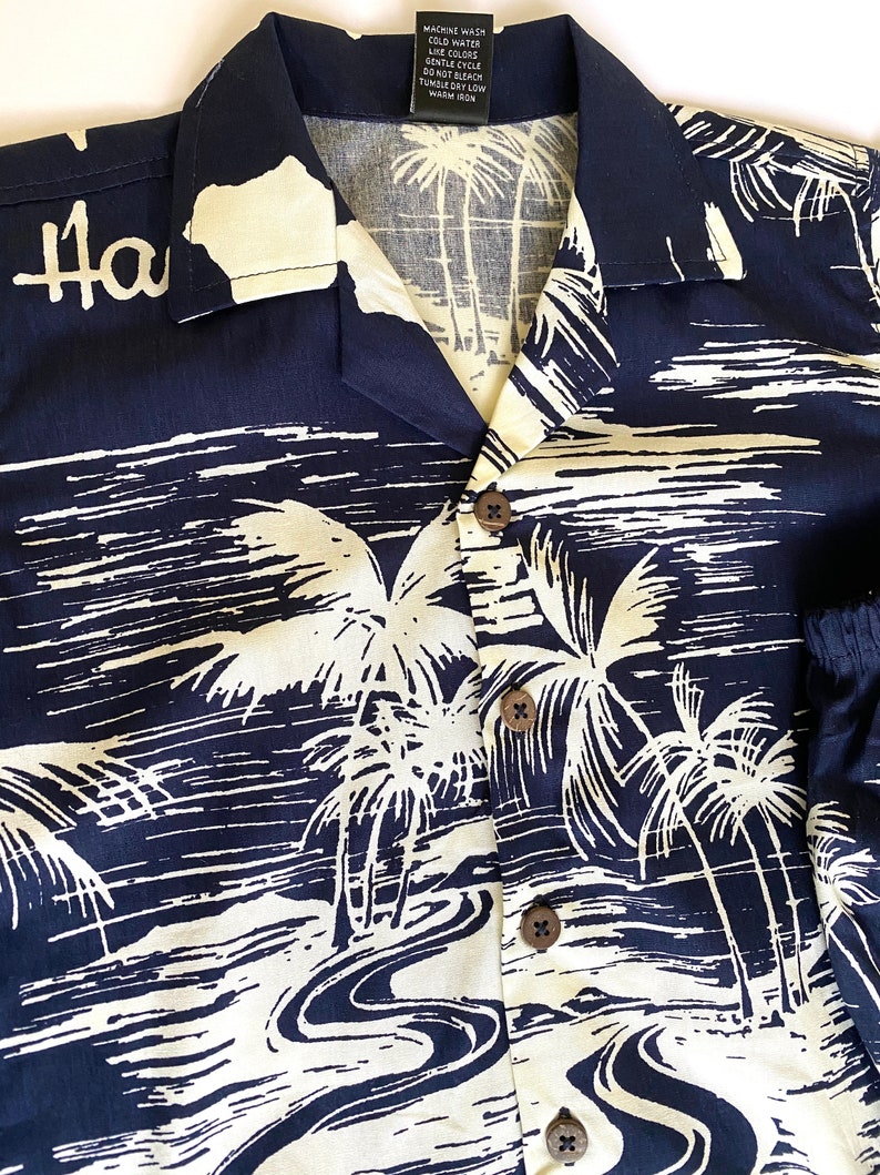 May include: A navy blue Hawaiian shirt with a white palm tree and river design. The shirt has a button-up front and a collar. The text "Ha" is visible on the left side of the shirt.