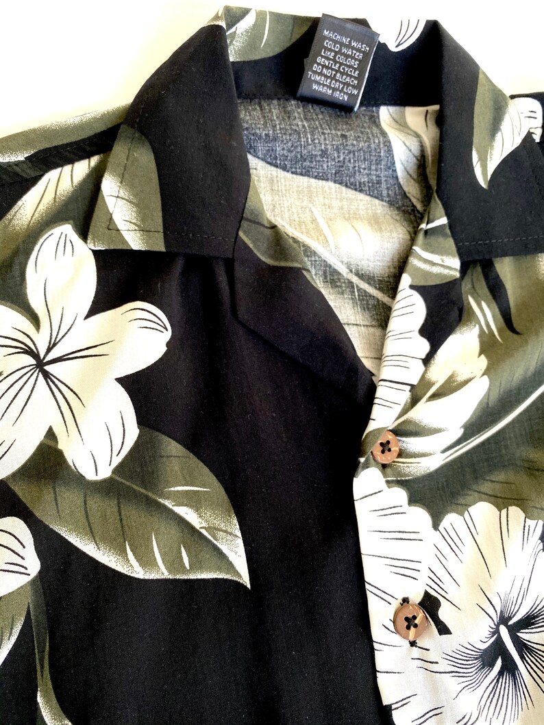 May include: Black and green Hawaiian shirt with white floral print. The shirt has a button-up collar and a care label that reads "Machine Wash Cold Water Like Colors Gentle Cycle Do Not Bleach Tumble Dry Low Warm Iron".