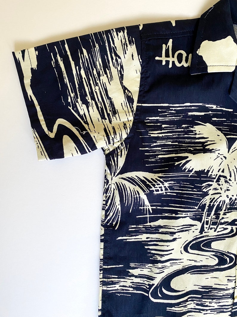 May include: A black and white short-sleeved button-up shirt with a palm tree and ocean scene. The shirt has the word "Hawaii" printed on the collar.