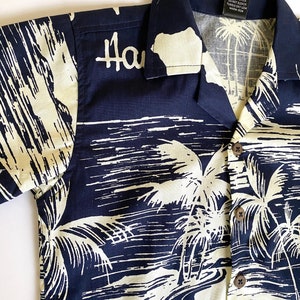 May include: A navy blue short-sleeved shirt with a white palm tree print. The shirt has a button-up collar and a pocket on the left side. The text "Hawaii" is printed on the front of the shirt.