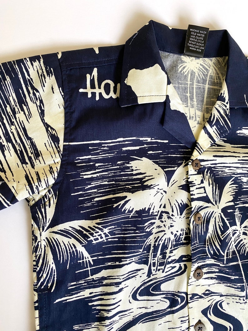 May include: A navy blue short-sleeved shirt with a white palm tree print. The shirt has a button-up collar and a pocket on the left side. The text "Hawaii" is printed on the front of the shirt.