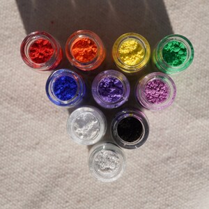 May include: Nine small glass jars filled with different coloured powders. The jars are arranged in a triangular shape on a white fabric surface. The colours are red, orange, yellow, green, blue, purple, white, black, and silver.