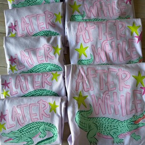 May include: Several light purple t-shirts with the phrases "LATER" and "AFTER WHILE" in pink lettering. Each shirt features a green alligator graphic, yellow stars, and pink star accents. The shirts are folded and stacked.