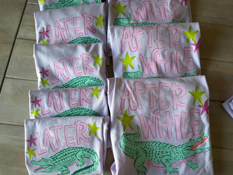 May include: Several light purple t-shirts with the phrases "LATER" and "AFTER WHILE" in pink lettering. Each shirt features a green alligator graphic, yellow stars, and pink star accents. The shirts are folded and stacked.