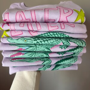 May include: A stack of lavender sweatshirts with pink text that reads "LATER" and a green alligator graphic. The sweatshirts are folded neatly, with the alligator design visible on the bottom layers. A yellow star is also visible.