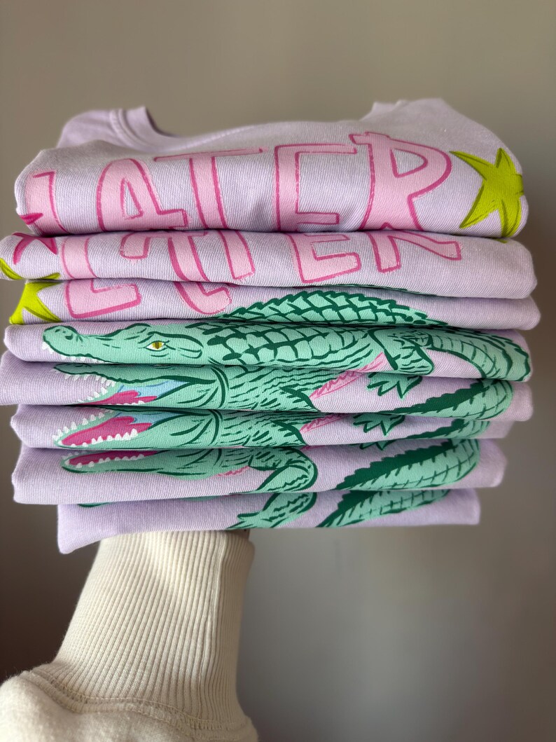 May include: A stack of lavender sweatshirts with pink text that reads "LATER" and a green alligator graphic. The sweatshirts are folded neatly, with the alligator design visible on the bottom layers. A yellow star is also visible.
