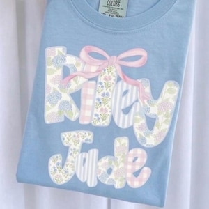 May include: A light blue t-shirt featuring the name Riley Jade printed in large, blocky letters. The letters are filled with floral, gingham, and striped patterns in shades of blue, pink, and green. A soft pink bow is tied over the name.