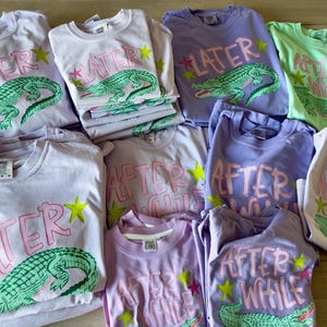 May include: A collection of light-colored t-shirts with graphic designs. The shirts feature text and an alligator illustration. The text on some shirts reads "LATER" and others read "AFTER WHILE". The alligator is green, and the text is pink.