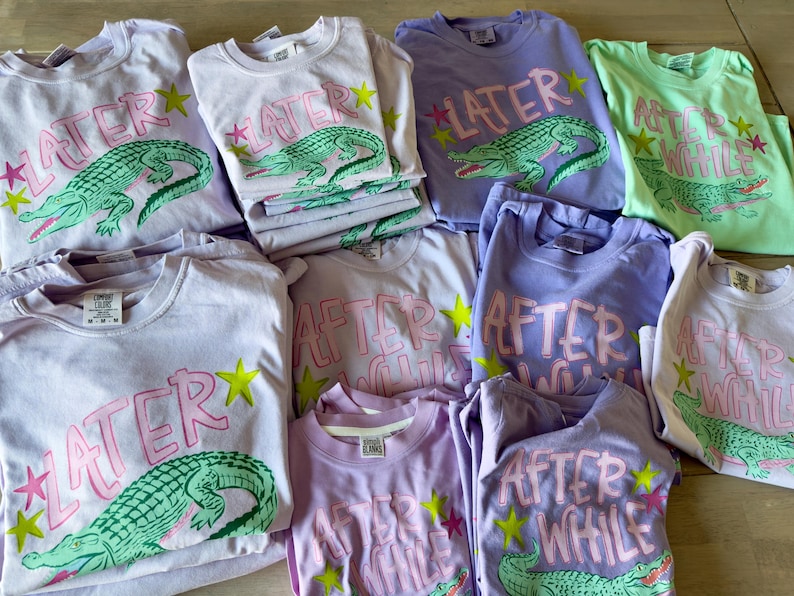 May include: A collection of light-colored t-shirts with graphic designs. The shirts feature text and an alligator illustration. The text on some shirts reads "LATER" and others read "AFTER WHILE". The alligator is green, and the text is pink.