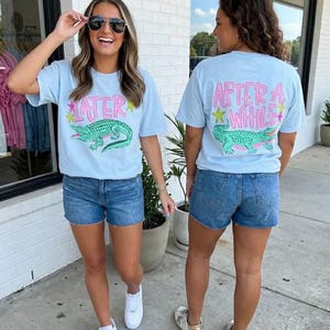 May include: Two people wearing light blue t-shirts and denim shorts. The front of the t-shirt has the word "LATER" above a green alligator graphic. The back of the t-shirt has the words "AFTER A WHILE" above the same alligator graphic.