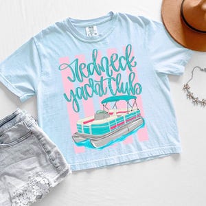 Pu&ograve; includere: Maglietta azzurra con la scritta "Redneck Yacht Club" in corsivo e una grafica di una barca pontone. L'immagine include pantaloncini di jeans, un cappello marrone e una collana floreale.