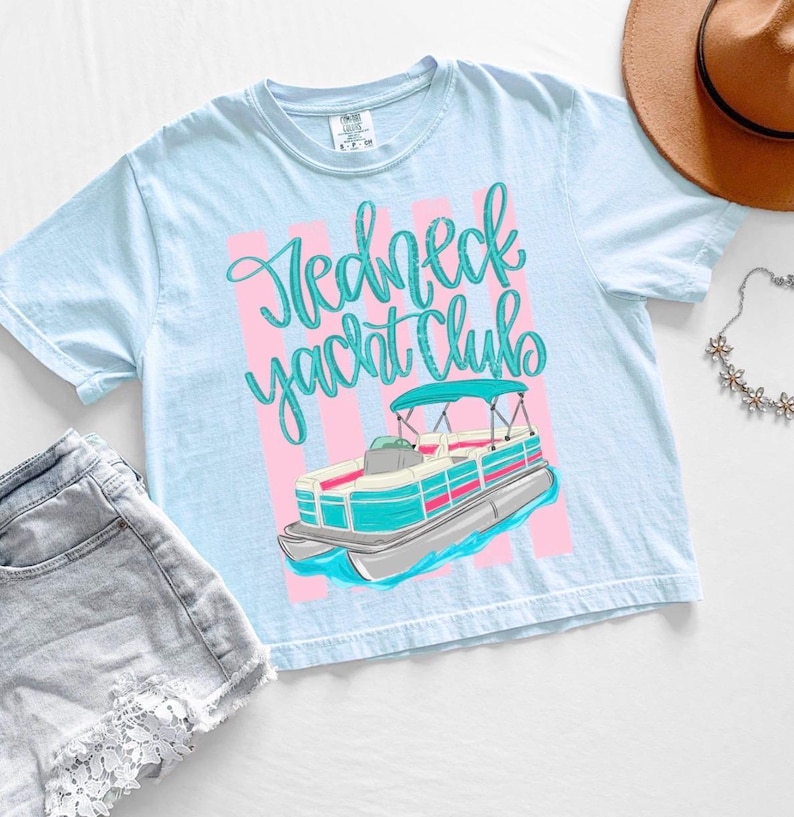 Pu&ograve; includere: Maglietta azzurra con la scritta "Redneck Yacht Club" in corsivo e una grafica di una barca pontone. L'immagine include pantaloncini di jeans, un cappello marrone e una collana floreale.