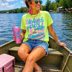 Pu&ograve; includere: Una donna in barca indossa una maglietta giallo neon con la scritta "Redneck Yacht Club" e una grafica di una barca. Indossa pantaloncini di jeans, occhiali da sole e accessori colorati. Tiene in mano un bicchiere glitterato rosa.