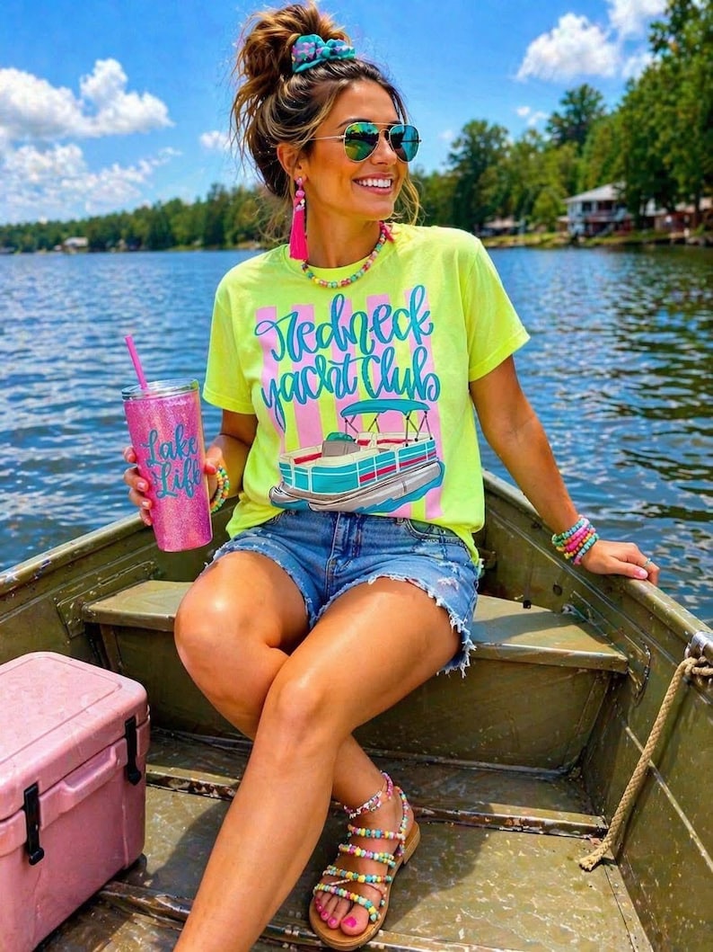 Pu&ograve; includere: Una donna in barca indossa una maglietta giallo neon con la scritta "Redneck Yacht Club" e una grafica di una barca. Indossa pantaloncini di jeans, occhiali da sole e accessori colorati. Tiene in mano un bicchiere glitterato rosa.