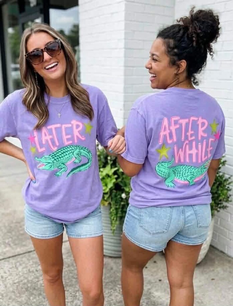 May include: Two women wearing matching purple t-shirts and denim shorts. The front of the shirts say "LATER" with a green alligator graphic. The back of the shirts say "AFTER WHILE" with a green alligator graphic.
