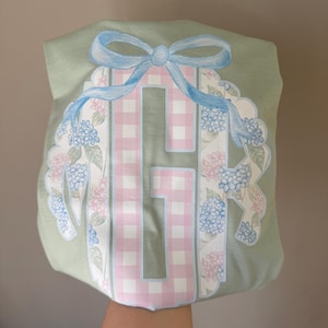 May include: A light green tote bag featuring a large letter "H" in a pink and white gingham pattern. The letter is surrounded by a floral wreath with blue and pink hydrangeas and a light blue bow at the top.