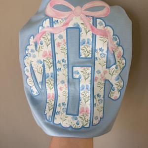 May include: A light blue sweatshirt featuring a floral monogram design. The monogram is filled with a pattern of pink and blue flowers. A pink bow is at the top of the monogram. The sweatshirt is held up by a person's arm.