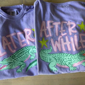 May include: Two lavender t-shirts with the words "LATER" and "AFTER WHILE" in pink letters. Each shirt features a green alligator graphic with an open mouth, pink accents, and yellow and pink star accents.