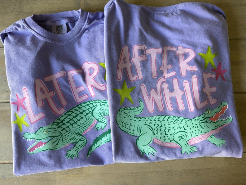 May include: Two lavender t-shirts with the words "LATER" and "AFTER WHILE" in pink letters. Each shirt features a green alligator graphic with an open mouth, pink accents, and yellow and pink star accents.