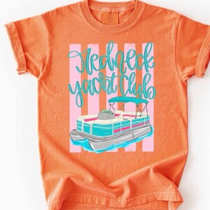 Pu&ograve; includere: T-shirt arancione con grafica di una barca pontone e la scritta "Redneck Yacht Club" in corsivo turchese. Il design della barca include dettagli turchesi, rosa e argento. Lo sfondo presenta strisce verticali rosa.