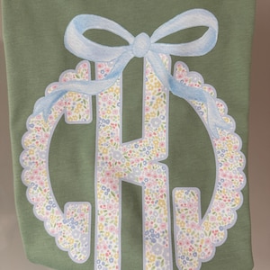 May include: Olive green t-shirt featuring a floral monogram with a light blue bow at the top. The monogram is outlined in light blue and filled with a pattern of small flowers in pastel colors. The bow adds a decorative touch to the design.