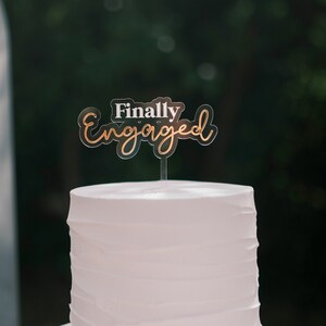 Könnte beinhalten: Eine zweistöckige weiße Hochzeitstorte mit einem Topper, auf dem "Endlich verlobt" steht.