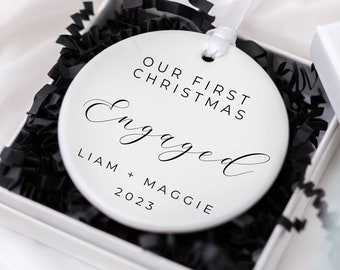May include: White ceramic Christmas ornament with the text "Our First Christmas Engaged Liam + Maggie 2023" in black lettering.