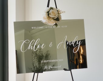 Puede incluir: Un espejo plateado con una inscripción blanca que dice "Bienvenido a la boda de Chloe & Andy, 22 de septiembre de 2023, The Ravenswood". El espejo está en un caballete negro con un pequeño ramo de flores blancas.
