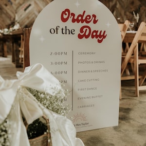 Op de afbeelding: Een wit trouwbord met de woorden "Order of the Day" in rood script. Het bord geeft het schema voor de huwelijksceremonie weer, inclusief tijd en evenement. Een mand met witte bloemen en een witte strik staat op de voorgrond.