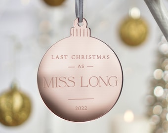 May include: A rose gold Christmas ornament with the text "LAST CHRISTMAS AS MISS LONG 2022" engraved on the front. The ornament is round with a loop for hanging and is suspended by a gray ribbon. Gold ornaments are visible in the background.