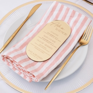 Könnte beinhalten: Eine formelle Tischdekoration mit einem weißen Teller, goldenem Besteck und einer rosa-weiß gestreiften Serviette. Eine goldene Menükarte mit dem Namen "Adam Raynor" und einem Menü liegt auf der Serviette.