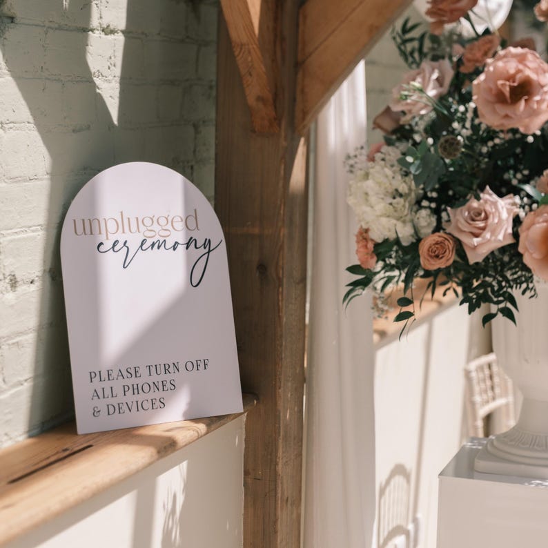 May include: A white sign with an arched top that reads "unplugged ceremony" in a script font. Below, it says "PLEASE TURN OFF ALL PHONES & DEVICES" in a sans-serif font. The sign is placed on a wooden shelf, with a floral arrangement in the background.